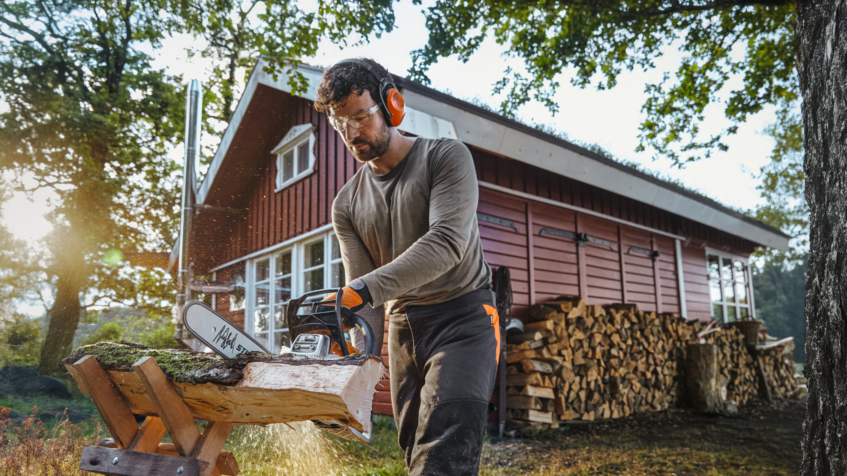 STIHL Kettensäge, mit der ein Mann einen Stamm vor einer roten Holzhütte mit einem Stapel Brennholz sägt